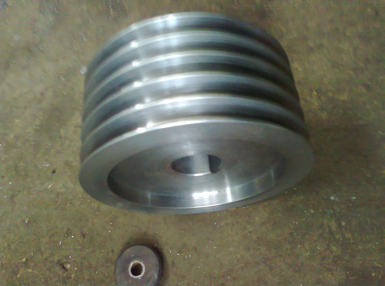 Metalowa, wielorowkowa rolka pasowa z otworem na klin, obok mały, okrągły element, leżąca na brudnej, metalowej powierzchni.