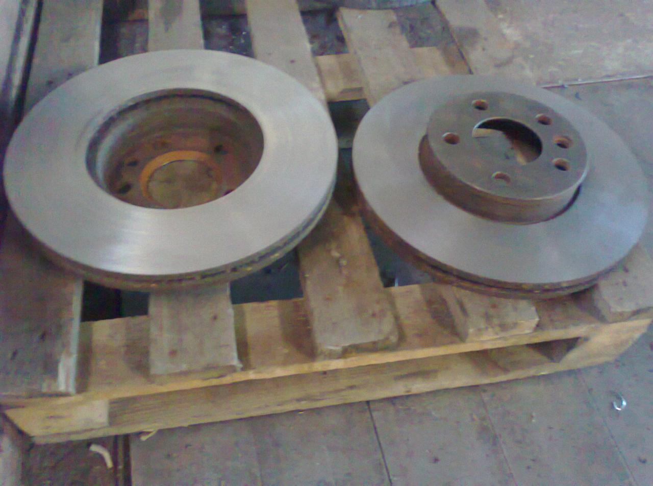 Dwa metalowe dyski hamulcowe, jeden z piastą z otworami na śruby, leżące na drewnianej palecie w warsztacie.