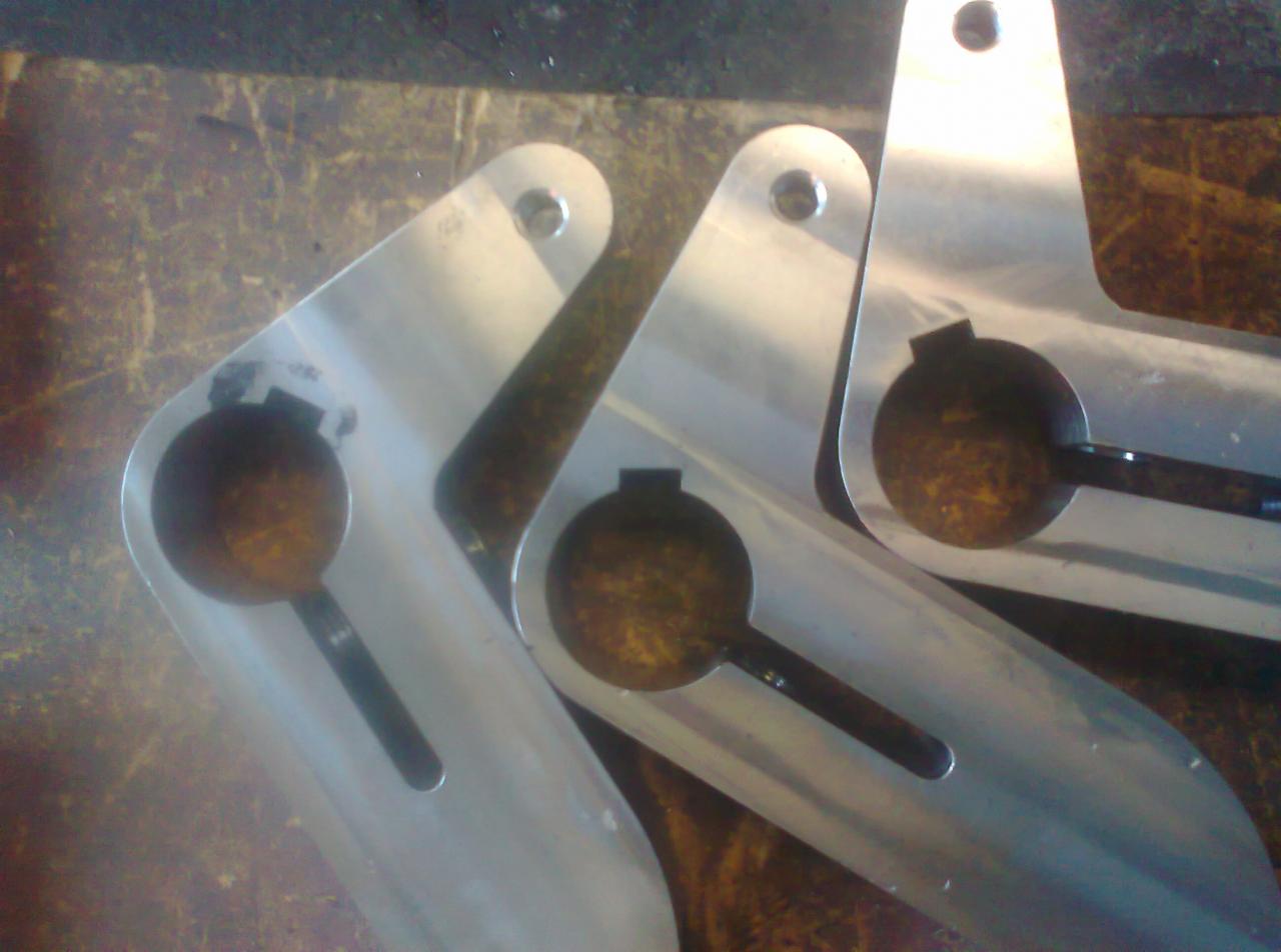 Trzy aluminiowe elementy z wyciętymi otworami i podłużnymi wgłębieniami, leżące na brudnej powierzchni.