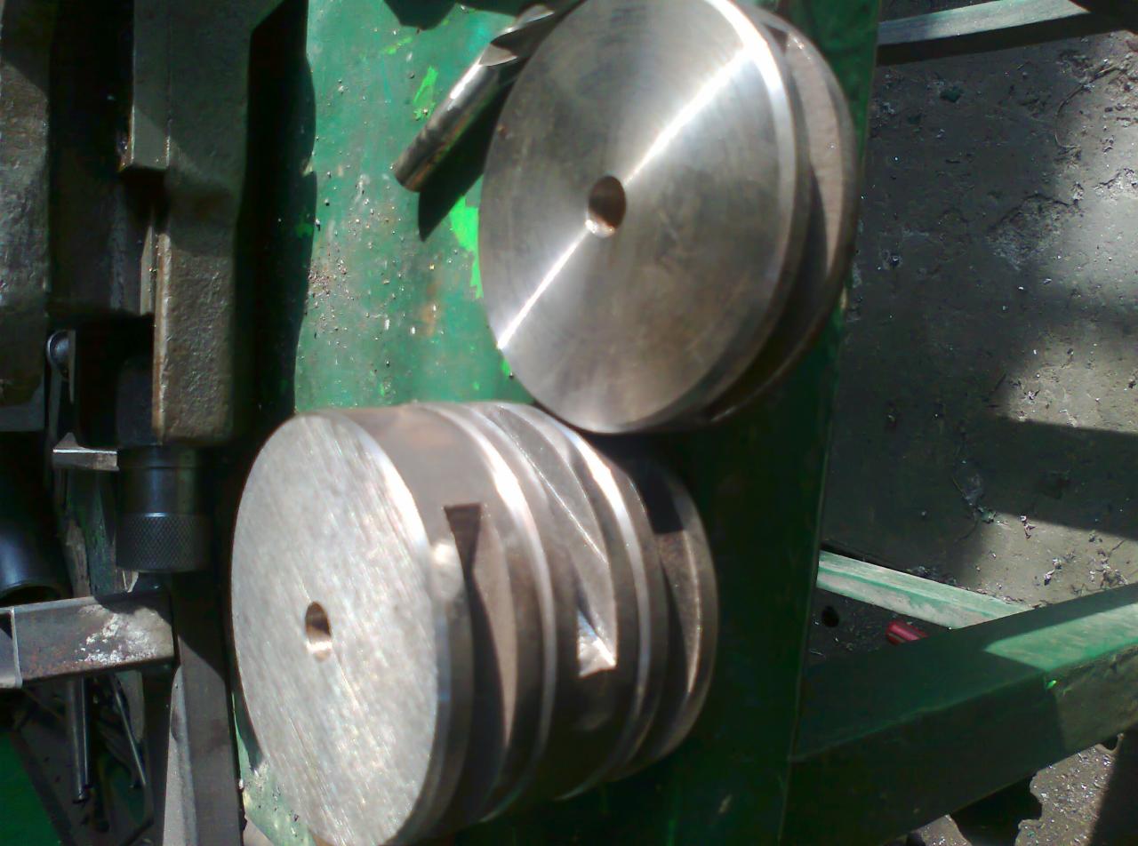 Dwa metalowe koła pasowe, jedno z rowkami, leżące na zielonej maszynie w warsztacie w Trzebnicy, w tle widoczne elementy tokarki.