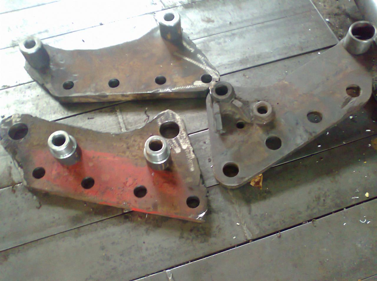 Trzy metalowe elementy konstrukcyjne, z widocznymi otworami i śladami obróbki, leżące na metalowej powierzchni warsztatowej.