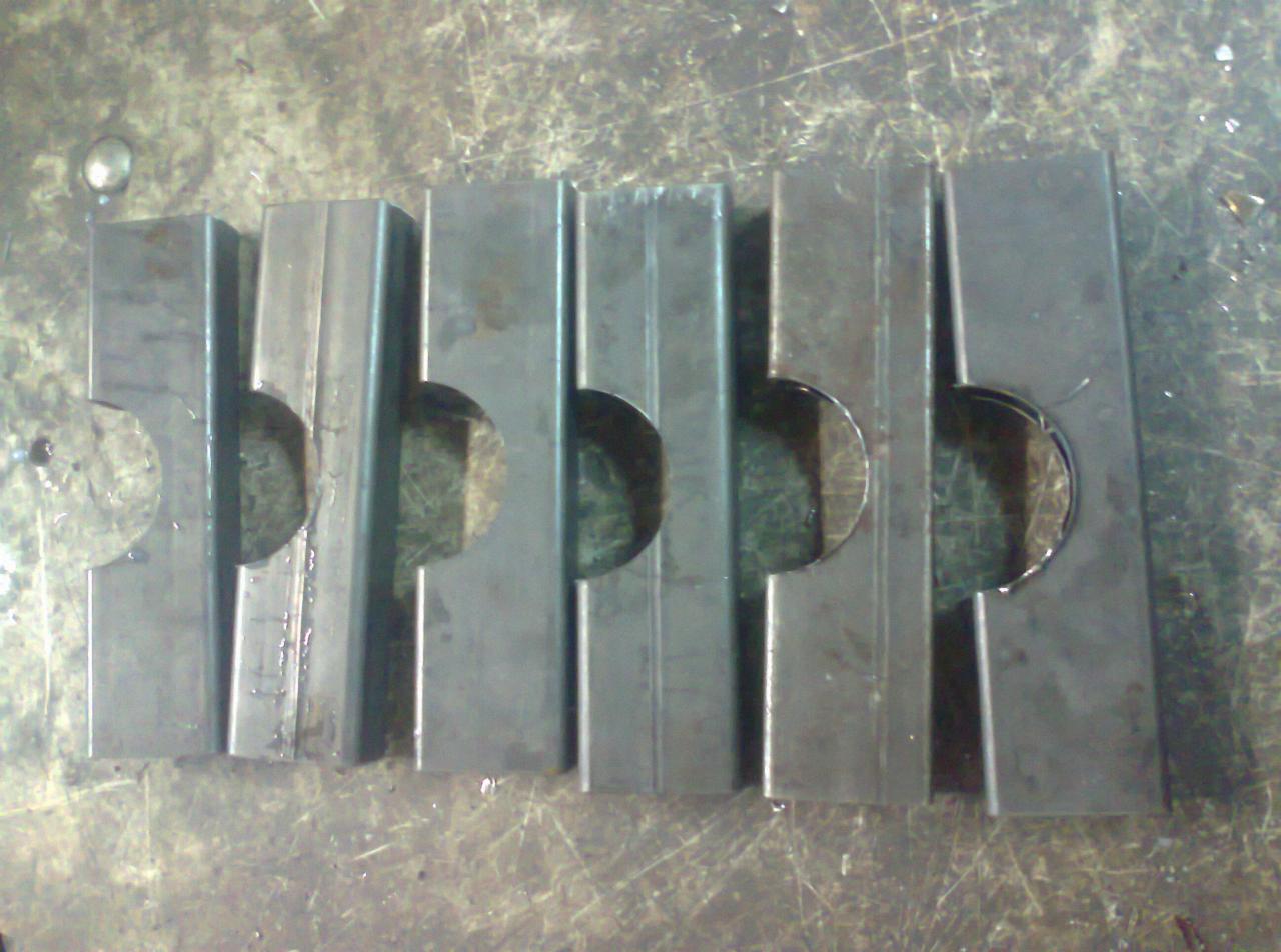 Szereg jednakowych, metalowych elementów z wyciętymi półokręgami i rowkami, leżących na brudnym, metalowym stole warsztatowym.