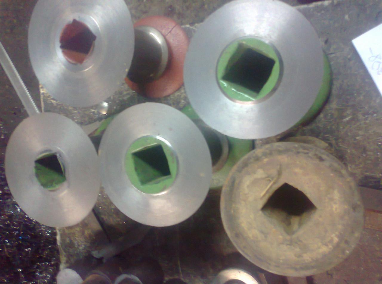 Metalowe pierścienie z kwadratowym otworem, jeden z nich skorodowany, pozostałe srebrne z zielonymi obwódkami, leżące na warsztatowym stole.