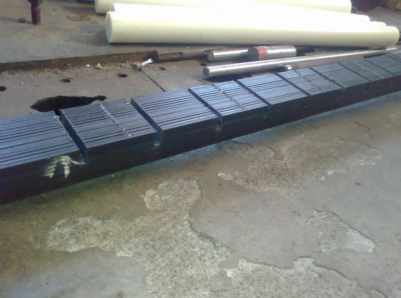 Czarny, stalowy profil z wyfrezowanymi rowkami, leżący na betonowej podłodze w warsztacie, z widocznymi białymi wałkami z tworzywa sztucznego w tle.