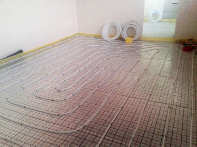 Białe rury grzewcze ułożone w pętle na metalowej siatce podłogowej, przygotowane do zalania wylewką, z rolkami rur w tle i żółtą taśmą zabezpieczającą przy ścianach.