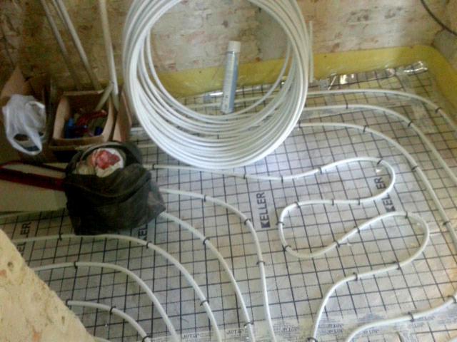 Białe rury ogrzewania podłogowego układane na izolacji z siatką, widoczny zwój rur i torba z narzędziami w tle.