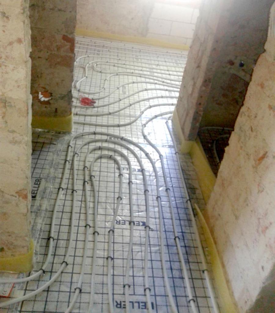 Montaż rur ogrzewania podłogowego na siatce, widoczne fragmenty ceglanych ścian i żółta izolacja przy ścianach.