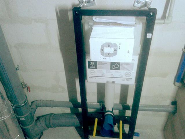 Stelaż podtynkowy WC zamontowany w surowym stanie, widoczne rury kanalizacyjne i przyłączeniowe, ściana w tle z widocznymi śladami po szpachlowaniu.