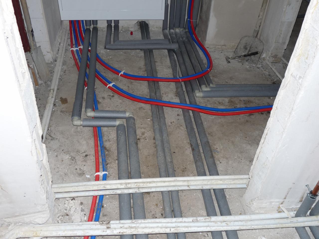 Rzut z góry na instalację wodną i kanalizacyjną w trakcie montażu, widoczne rury w izolacji oraz rury czerwone i niebieskie mocowane do podłoża, ściany w stanie surowym.