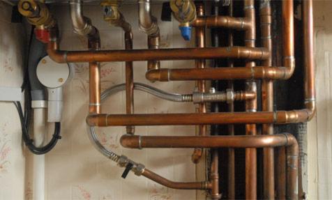 Skomplikowany układ miedzianych rur grzewczych z zaworami i elastycznymi przewodami, zamontowany na ścianie z tapetą w kwiatowy wzór.