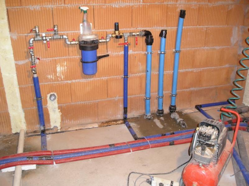 Instalacja wodna z niebieskich rur prowadzona w surowym stanie, widoczny filtr wody, kompresor i fragment ściany z cegły dziurawki.