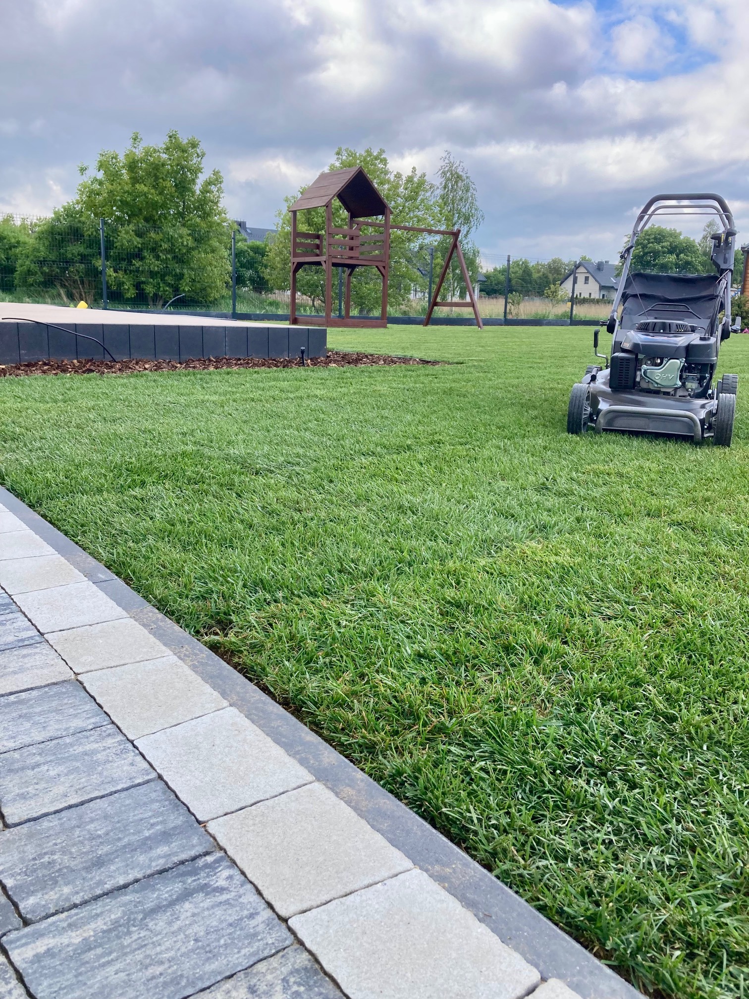 Świeżo skoszony trawnik w Bożej Woli z widoczną kosiarką, placem zabaw i wykończonym obrzeżem z kostki brukowej. Trawa jest gęsta i zielona, a na dalszym planie widoczne są drzewa i zabudowania.