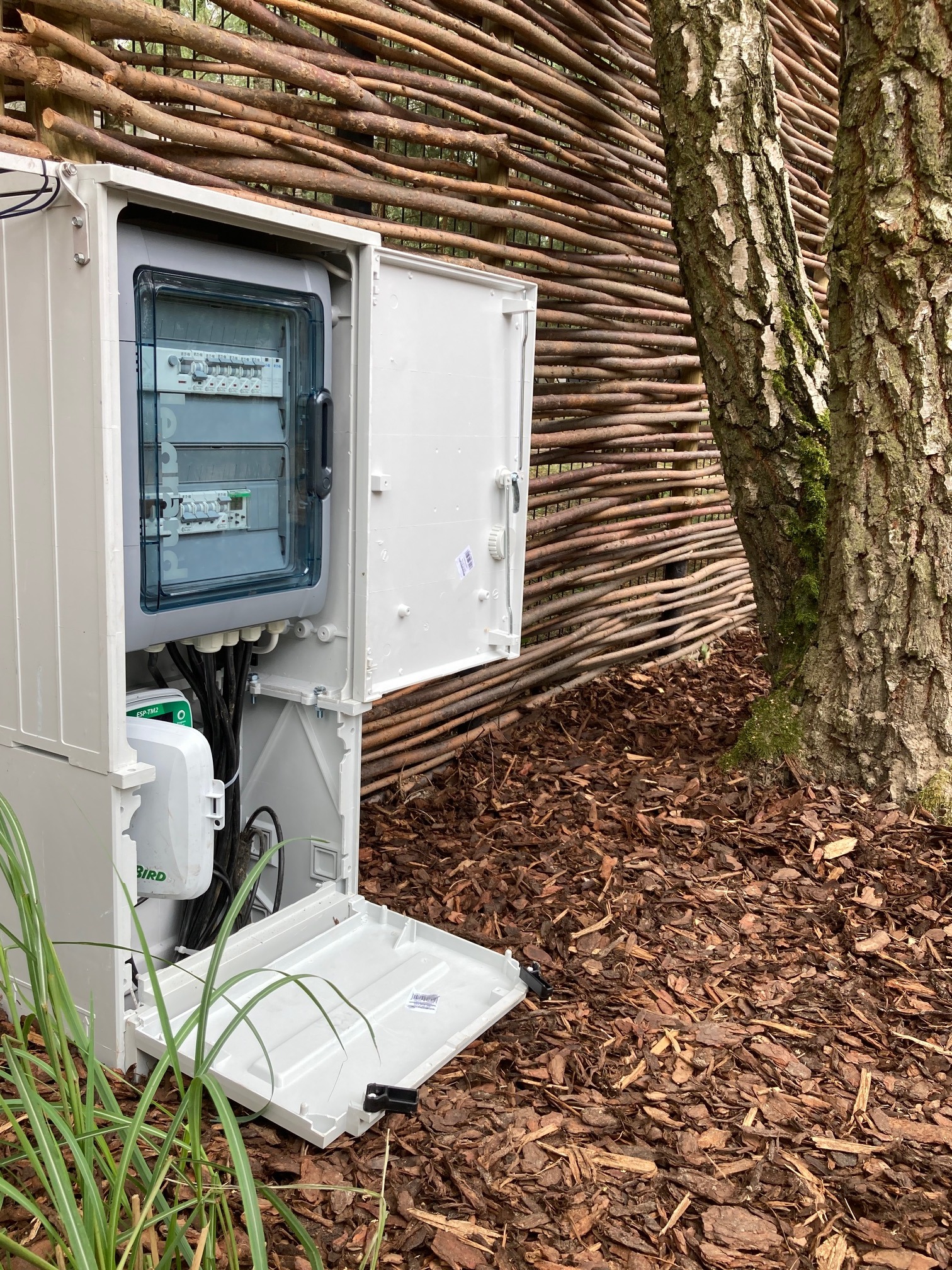Otwarta szafka sterownicza systemu nawadniania z widocznymi przewodami i elementami elektrycznymi, umieszczona na korze drzewnej obok pnia drzewa i plecionej ściany z gałęzi.