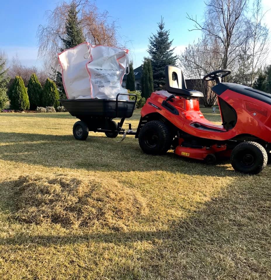 Czerwony traktorek ogrodowy z przyczepką wypełnioną workiem na trawie po wertykulacji, drzewa iglaste w tle.