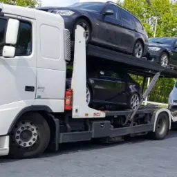 Ciężarówka Volvo FH z naczepą transportującą cztery samochody osobowe różnych marek, zaparkowana na parkingu z widocznym lasem w tle.
