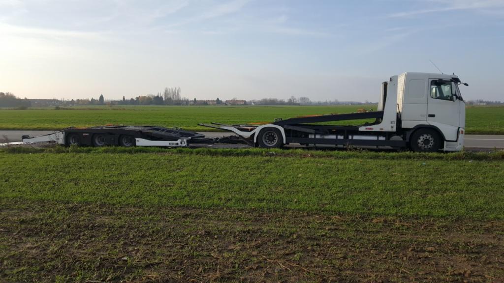 Ciężarówka z platformą transportową stoi na poboczu drogi w Pleszewie, widok na zielone pole i odległe zabudowania w tle.