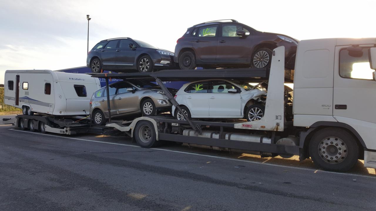 Laweta przewozi uszkodzony biały samochód osobowy oraz trzy inne pojazdy, w tym przyczepę kempingową Hobby, na dwupoziomowej platformie transportowej.