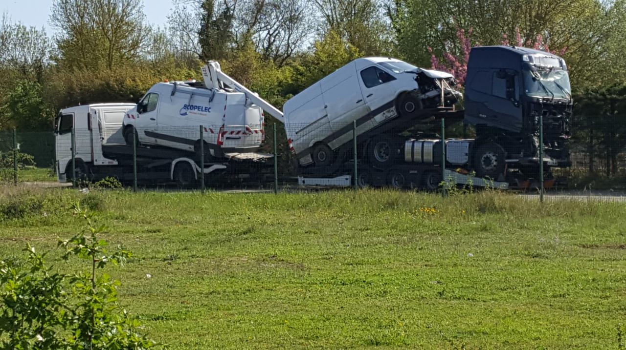 Dwa samochody ciężarowe przewożące uszkodzone furgonetki, widoczne uszkodzenia karoserii, ujęcie zza ogrodzenia z zielonym polem na pierwszym planie.