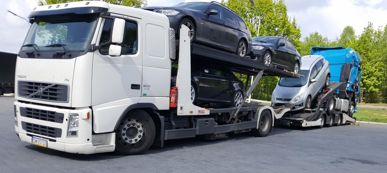 Ciężarówka Volvo FH z naczepą transportującą cztery samochody osobowe różnych marek, zaparkowana na parkingu z widocznym lasem w tle.
