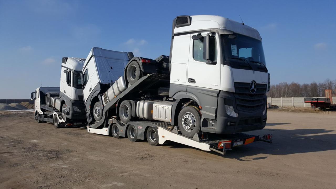 Dwa białe samochody ciężarowe Mercedes-Benz Actros na niskopodwoziowej lawecie, widoczne tablice rejestracyjne, w tle plac budowy i błękitne niebo.