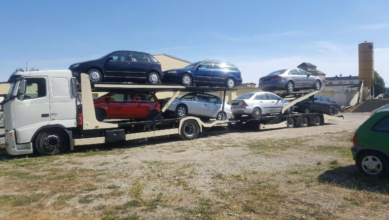 Laweta z białym ciągnikiem Volvo przewozi sześć samochodów osobowych różnych marek i kolorów na dwupoziomowej platformie transportowej. W tle widoczny budynek przemysłowy i błękitne niebo.