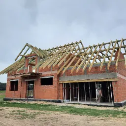 Surowy stan budynku jednorodzinnego z czerwonej cegły, z widoczną konstrukcją drewnianą dachu i metalowymi podporami, na tle pochmurnego nieba.