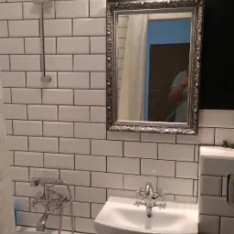 Wykończona łazienka z białą armaturą, baterią retro, lustrem w ozdobnej srebrnej ramie i białymi płytkami typu metro, z ciemną ścianą w tle.