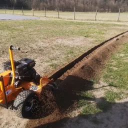 Pomarańczowa glebogryzarka wykopuje wąski rów w ziemi na trawiastym terenie, widoczny długi wykop z usypaną ziemią, w tle drzewa i ogrodzenie.
