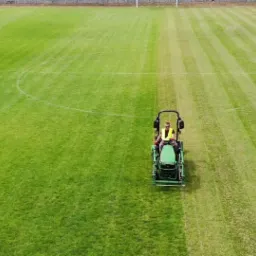 Operator kosiarki traktorowej w żółtej kamizelce kosi trawę na boisku sportowym, widok z góry ukazuje różnicę w wysokości trawy przed i po koszeniu.
