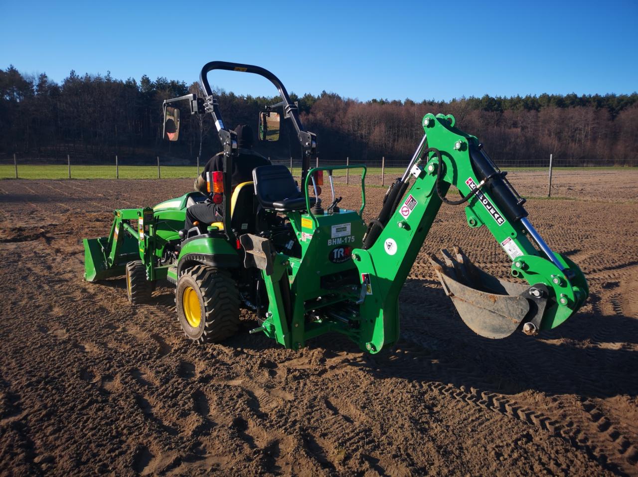 Zielony traktor z łyżką kopiącą na polu z widocznymi śladami opon, w tle drzewa pod błękitnym niebem.