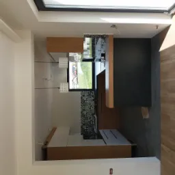 Wnętrze kuchni w trakcie wykańczania: widoczne gładkie, białe ściany, drewniane elementy zabudowy, czarna ściana oraz fragment podłogi z płytek imitujących drewno, z widocznymi przewodami...