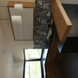 Nowoczesna kuchnia z białymi szafkami, drewnianymi elementami i czarno-białymi płytkami dekoracyjnymi na ścianie, widok z dołu na sufit i okno.