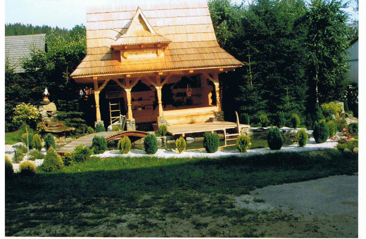 Altana w stylu góralskim z drewnianym dachem krytym gontem, wsparta na kamienno-drewnianych kolumnach, z małym mostkiem i oczkiem wodnym w otoczeniu zieleni.