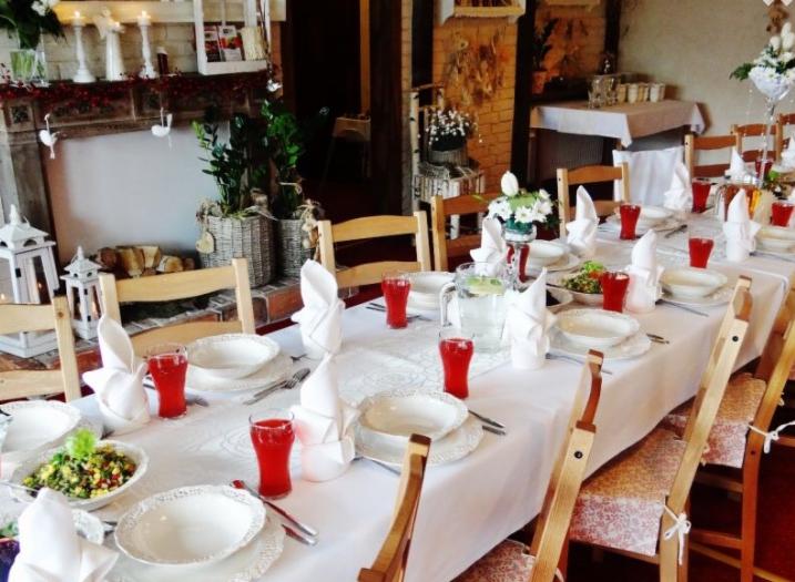 Uroczysty stół nakryty białym obrusem, z białymi talerzami, serwetkami ułożonymi w piramidy, szklankami z czerwonym napojem i miseczkami z sałatką, ustawiony w przestronnym wnętrzu z dekoracjami.