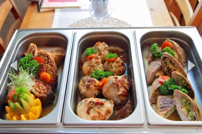 Metalowa taca z trzema przegródkami wypełniona różnorodnymi daniami mięsnymi: roladki, pieczenie, dekorowane zieleniną i pomarańczą.