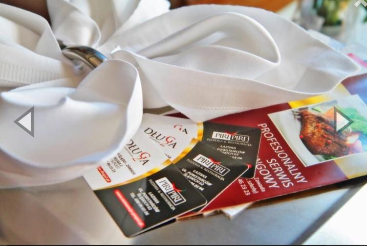 Elegancko złożona, biała serwetka z metalowym pierścieniem, leżąca na ulotkach reklamowych pizzerii i restauracji z Lazisk Górnych, w tle rozmyte elementy wystroju.