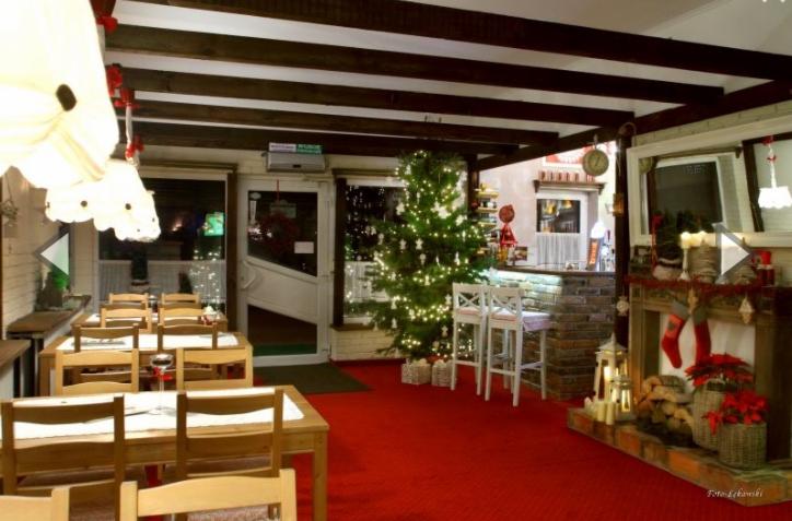 Uroczysta sala restauracyjna z czerwonym dywanem, drewnianymi stołami i krzesłami, barem z kamienia, kominkiem z dekoracjami świątecznymi i choinką z lampkami.