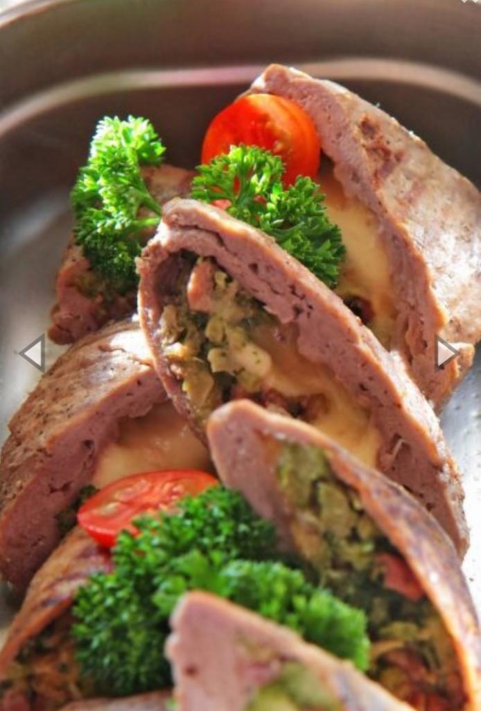 Krojony mięsny rolad z farszem warzywnym i serem, udekorowany natką pietruszki i pomidorem, podany na metalowej tacy.