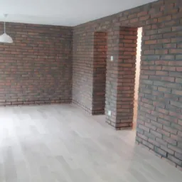 Salon z ceglaną ścianą, jasnymi panelami podłogowymi i telewizorem na ścianie. Widoczne drzwi bez futryny również wykończone cegłą.