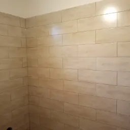 Wykończona ściana w łazience z beżowymi, podłużnymi płytkami imitującymi drewno, widoczny otwór w ścianie na dole po lewej stronie i fragment podłogi z ciemnymi plamami.