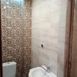 Wykończona łazienka z toaletą i umywalką, ściany wyłożone płytkami w dwóch wzorach: beżowymi imitującymi marmur i brązowymi z ornamentami, widoczna kratka wentylacyjna i włącznik światła.