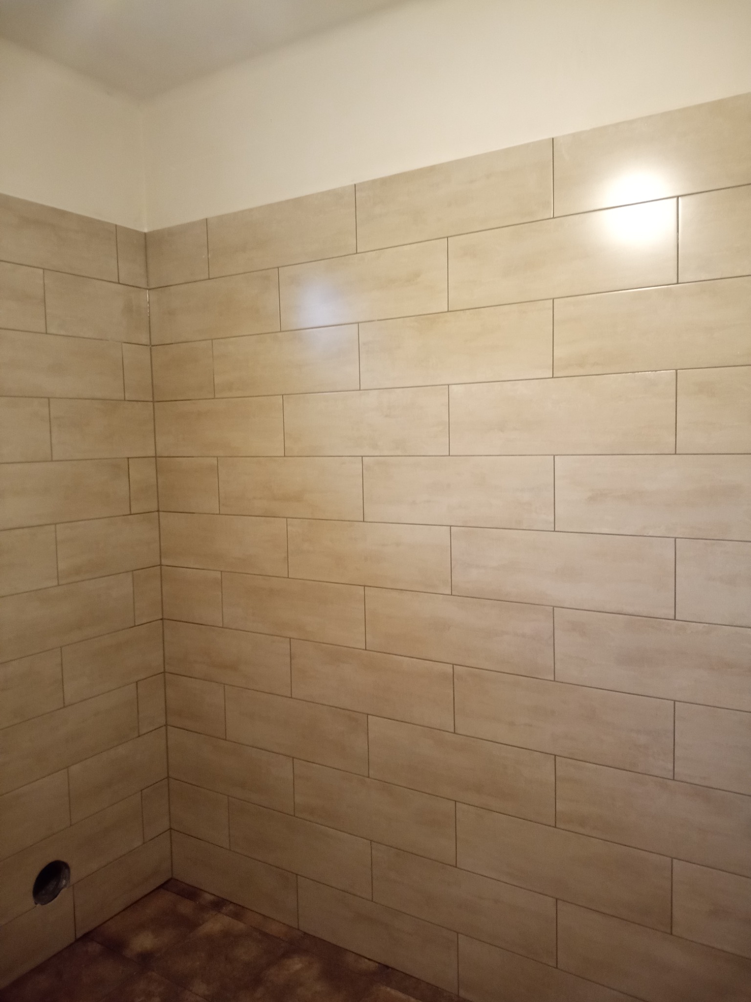 Wykończona ściana w łazience z beżowymi, podłużnymi płytkami imitującymi drewno, widoczny otwór w ścianie na dole po lewej stronie i fragment podłogi z ciemnymi plamami.