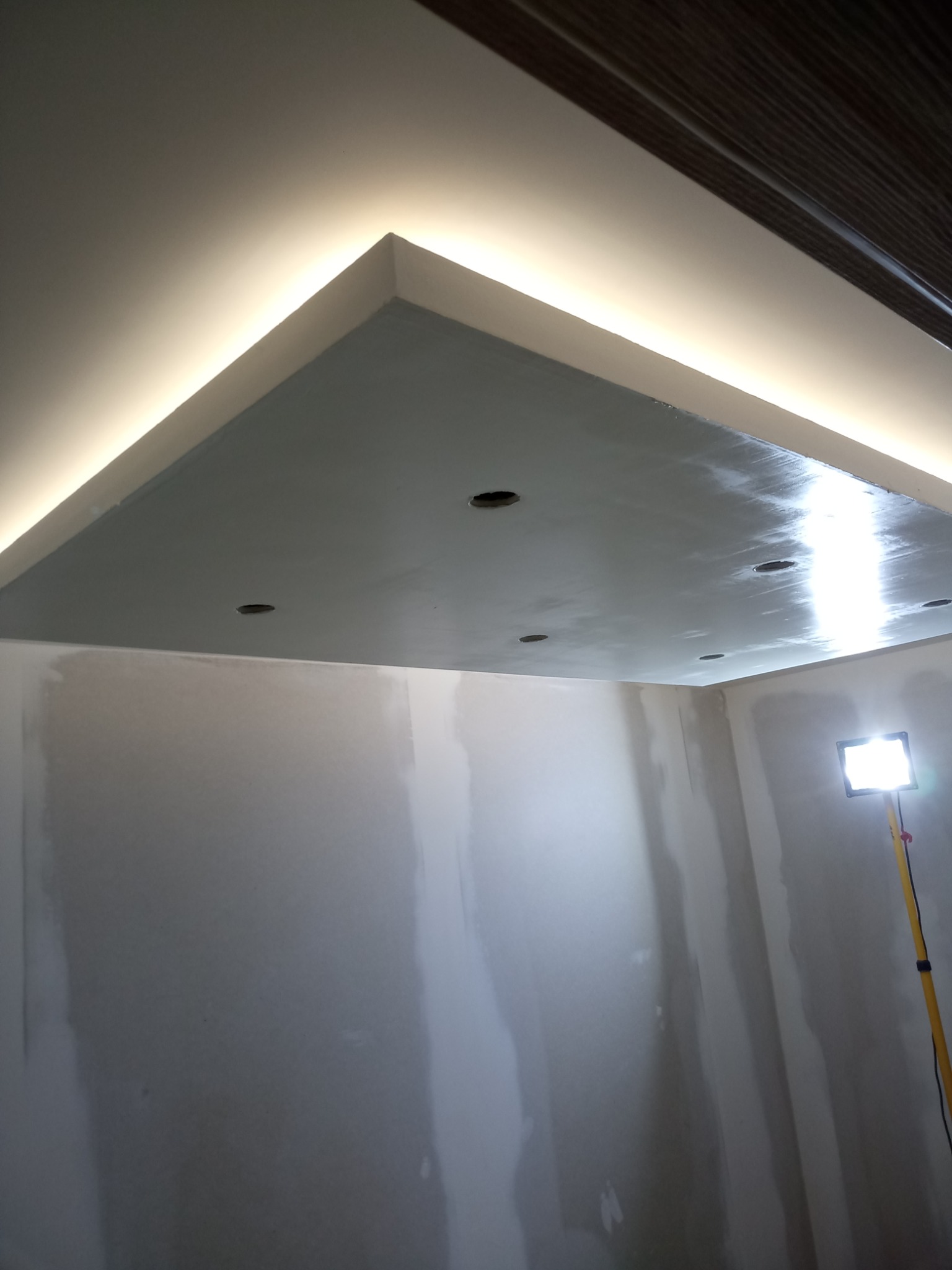 Sufit podwieszany z oświetleniem LED w trakcie montażu, widoczne otwory na oświetlenie punktowe i niedokończone ściany gipsowe, oświetlone lampą roboczą.