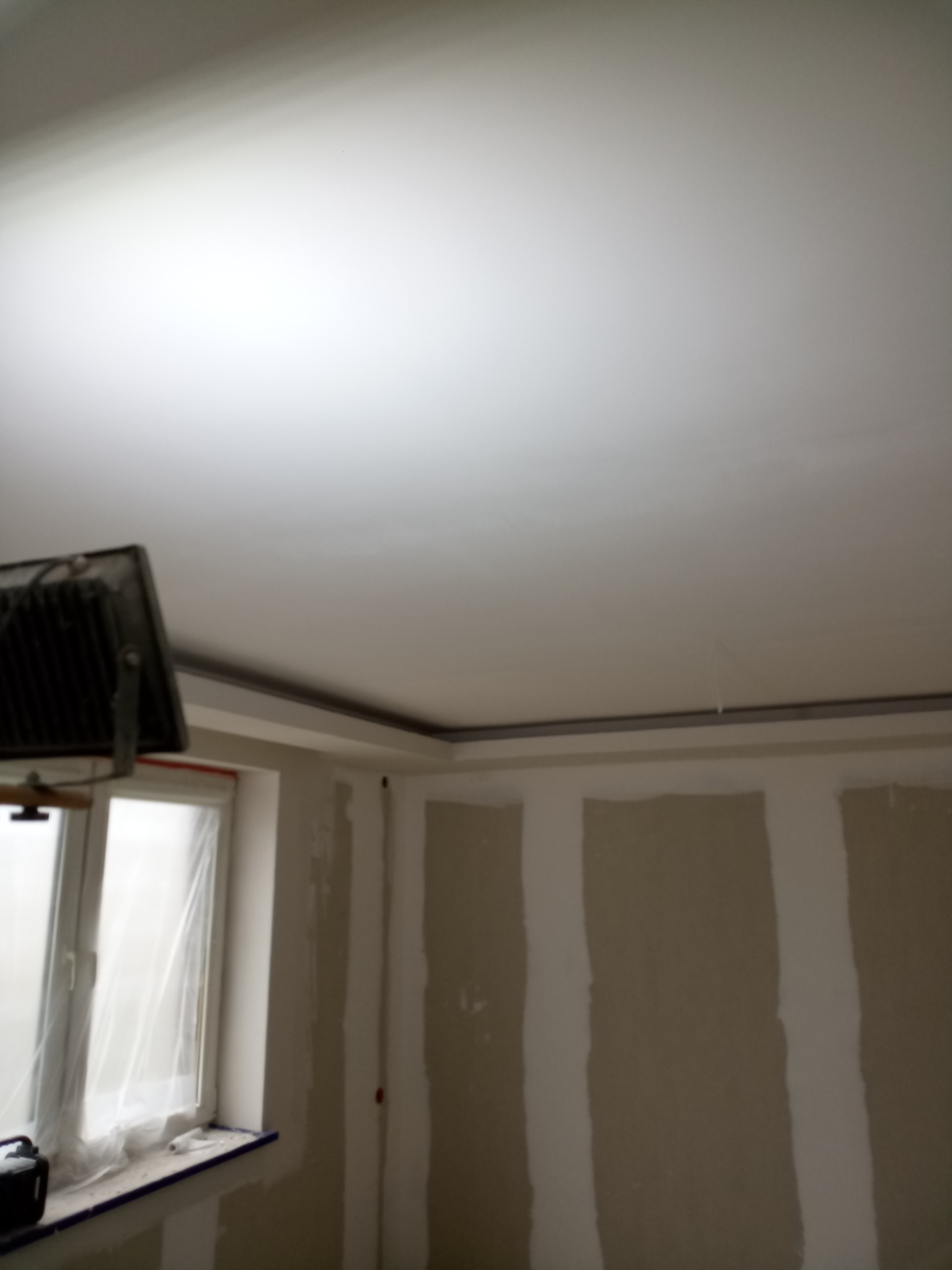 Pokój w trakcie remontu: ściany pokryte płytami gipsowo-kartonowymi, sufit pomalowany na biało, widoczne oświetlenie robocze i okno zabezpieczone folią.
