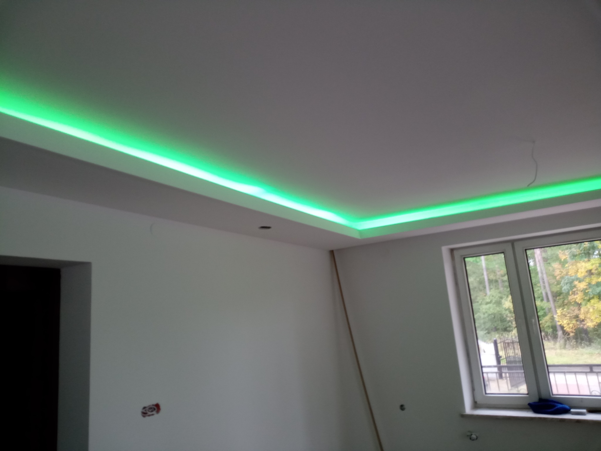 Białe wnętrze pokoju z podwieszanym sufitem i zielonym oświetleniem LED, widok na okno z widokiem na drzewa, ściany w trakcie prac wykończeniowych.