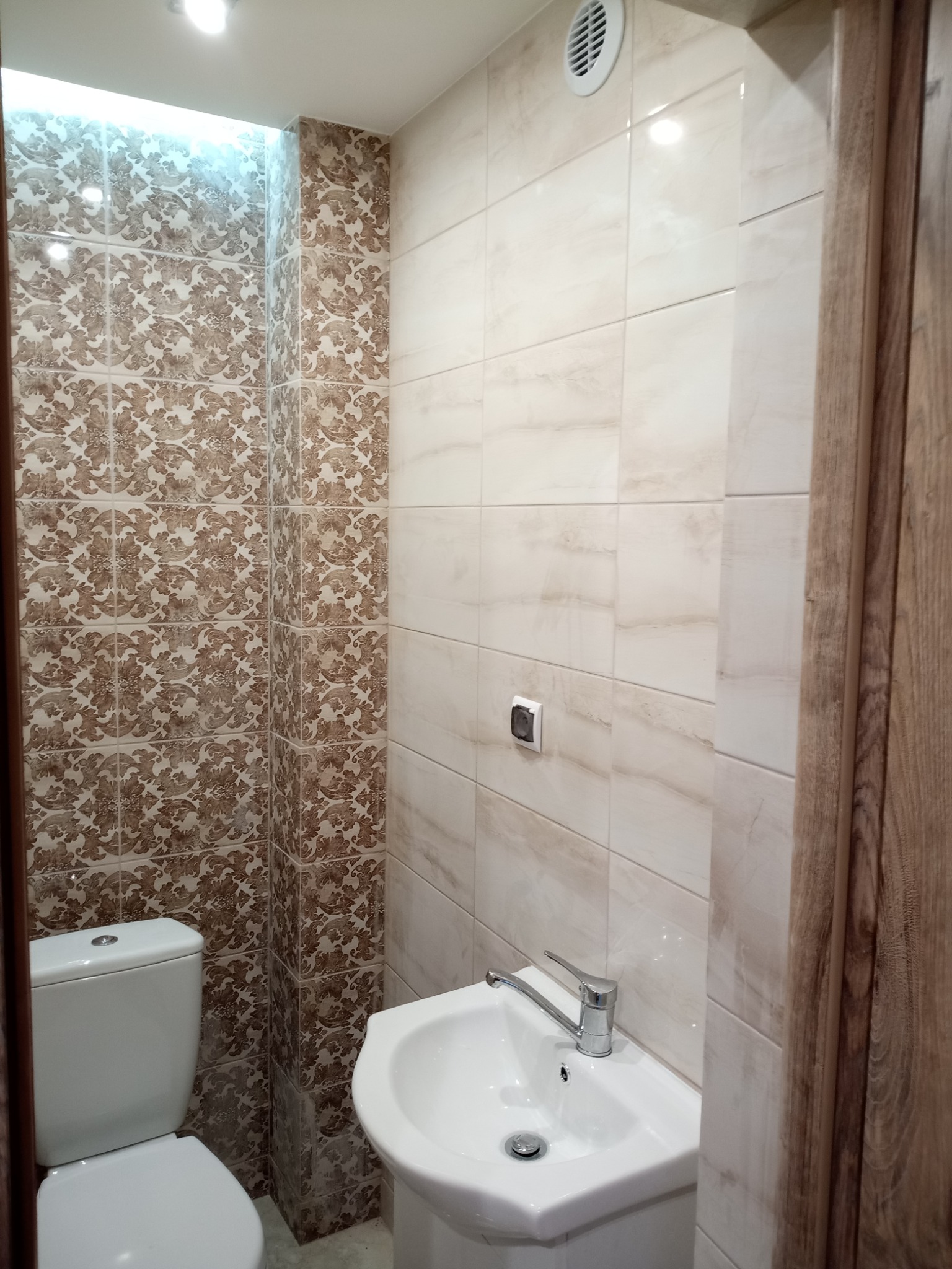 Wykończona łazienka z toaletą i umywalką, ściany wyłożone płytkami w dwóch wzorach: beżowymi imitującymi marmur i brązowymi z ornamentami, widoczna kratka wentylacyjna i włącznik światła.