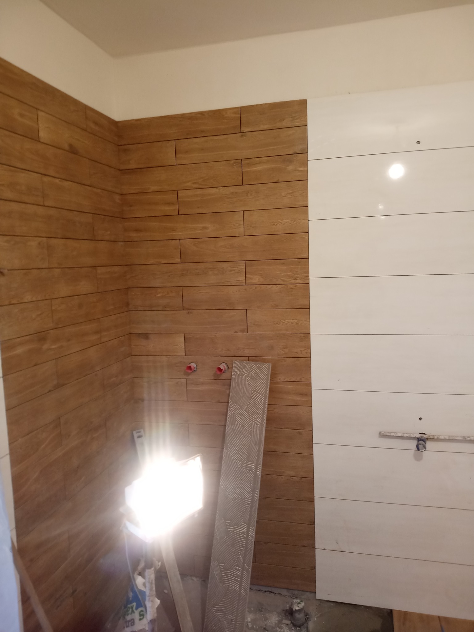 Ściana łazienki wyłożona częściowo brązowymi płytkami imitującymi drewno, a częściowo białymi płytkami w poziome pasy, z widocznymi rurami i lampą budowlaną.