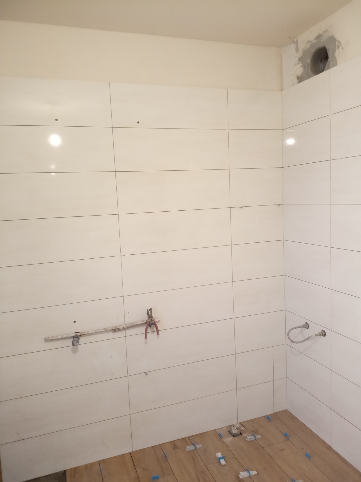 Ściany łazienki w trakcie układania białych płytek, widoczne narzędzia hydrauliczne, niezabezpieczony otwór wentylacyjny i fragment podłogi z drewnopodobnych paneli z niebieskimi krzyżykami...