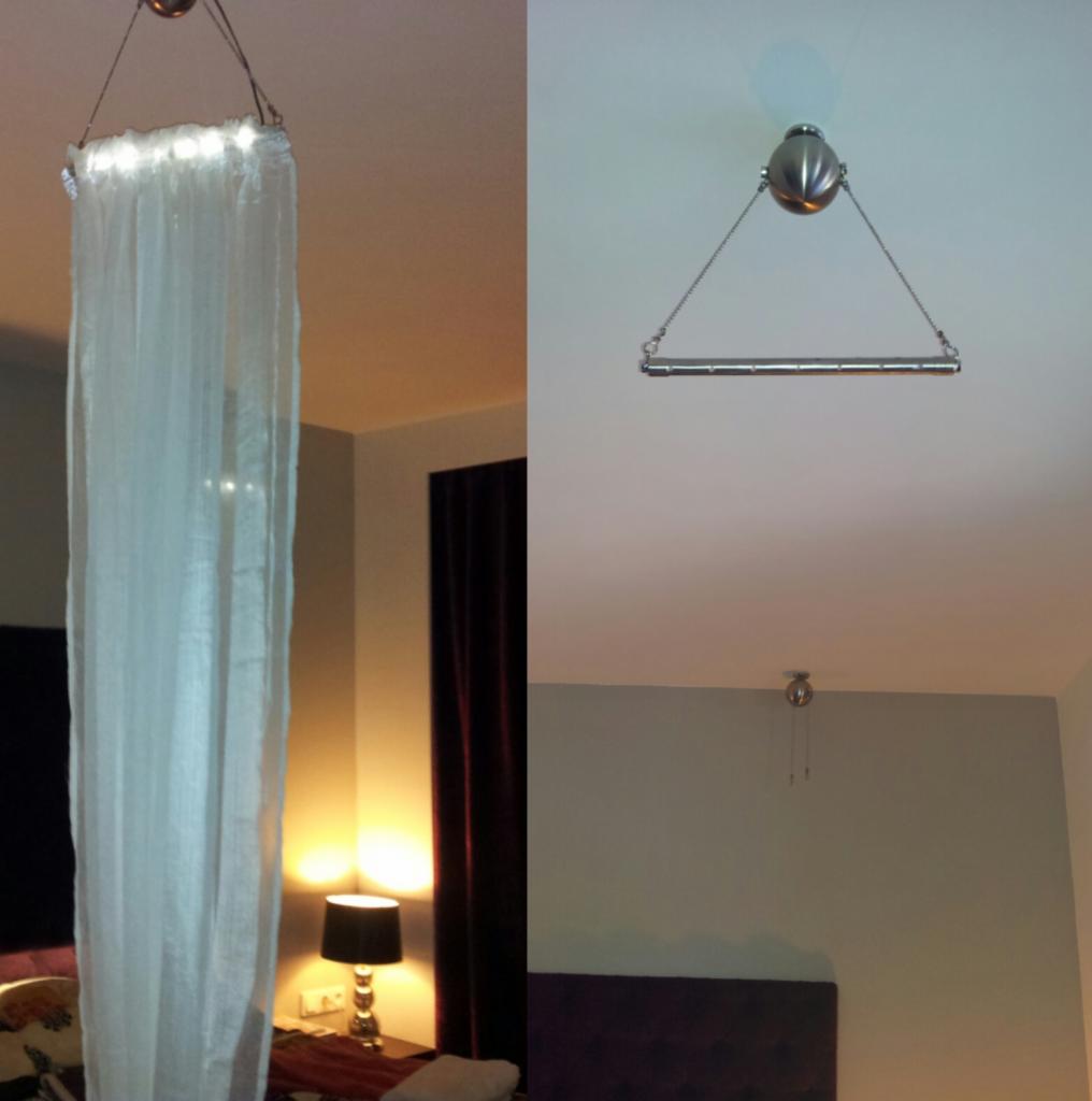 Sypialnia z baldachimem wykonanym z białej, prześwitującej tkaniny, podwieszonym na metalowym stelażu z łańcuszkami, oświetlonym od góry, widocznym na tle ściany i lampki nocnej.