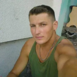 Młody mężczyzna w zielonym podkoszulku i srebrnym łańcuszku pozuje na tle szarej ściany z zielonym akcentem.